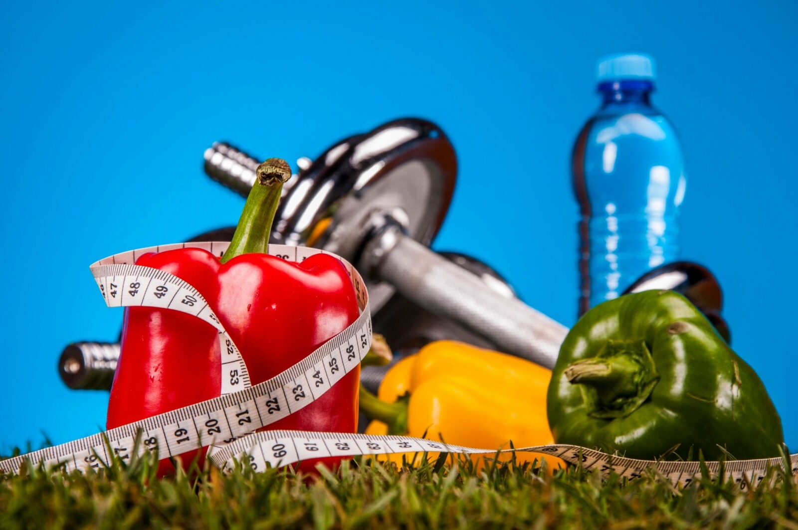 Three bell peppers with a barbell and a water bottle outside in the grass