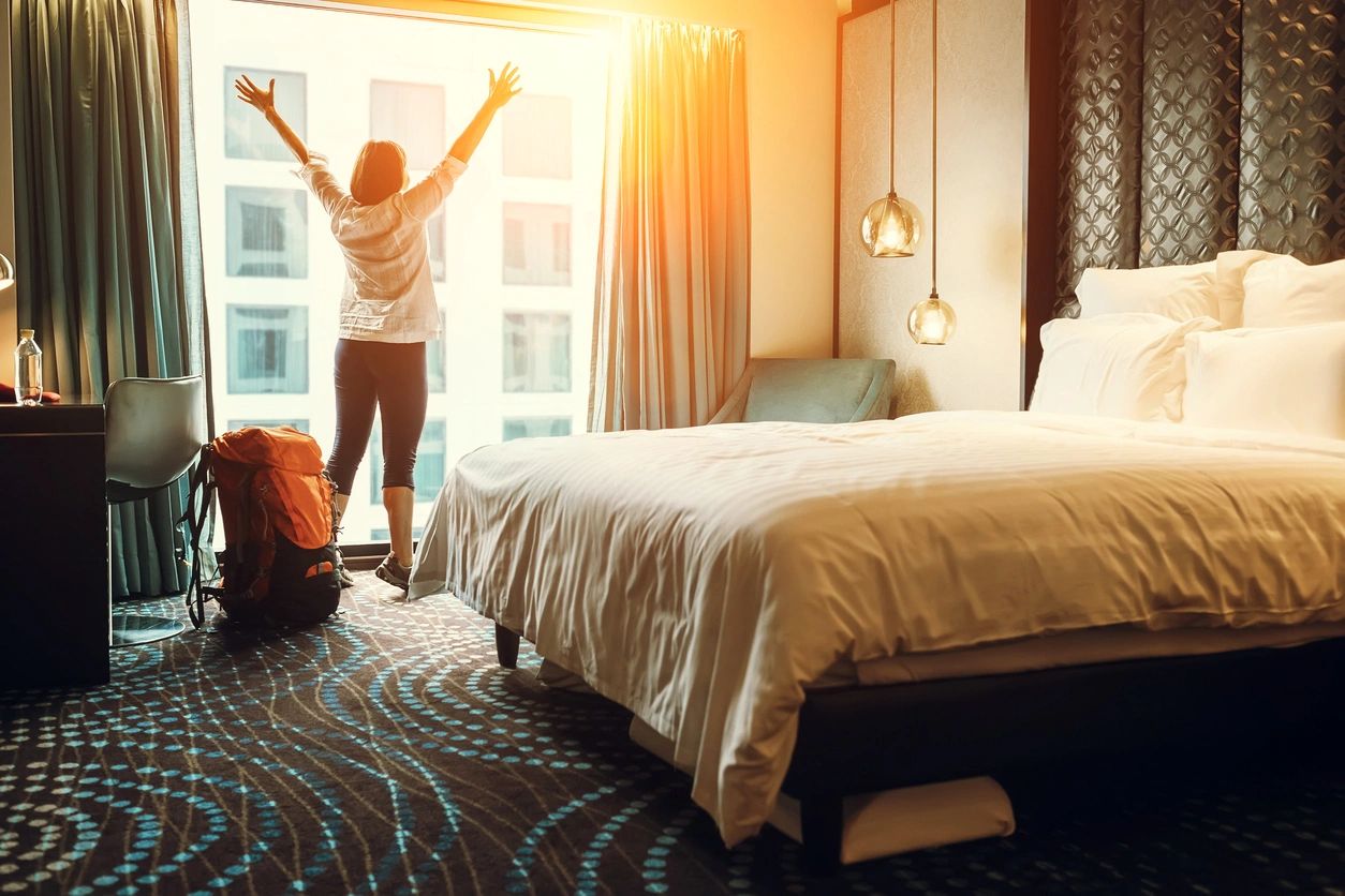 Woman in a hotel room with large bed and the sun beating in the window.